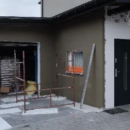 Elewacja budynku w trakcie prac: nałożony tynk w odcieniach szarości, widoczne rusztowania, worki z materiałem w garażu, okno zabezpieczone folią, przygotowane wiadra z zaprawą, częściowo ocieplone...