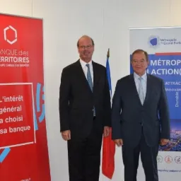 Dwaj mężczyźni w garniturach stoją przed banerami Banque des Territoires i Métropole du Grand Paris, z flagą Francji pośrodku.