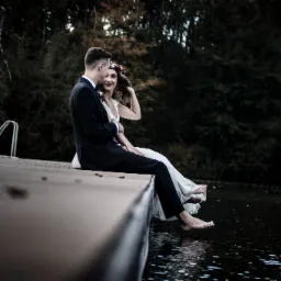 Para młoda siedzi na drewnianym pomoście nad wodą, boso, ona w wianku na głowie, on w czarnym garniturze, w tle ciemny las.