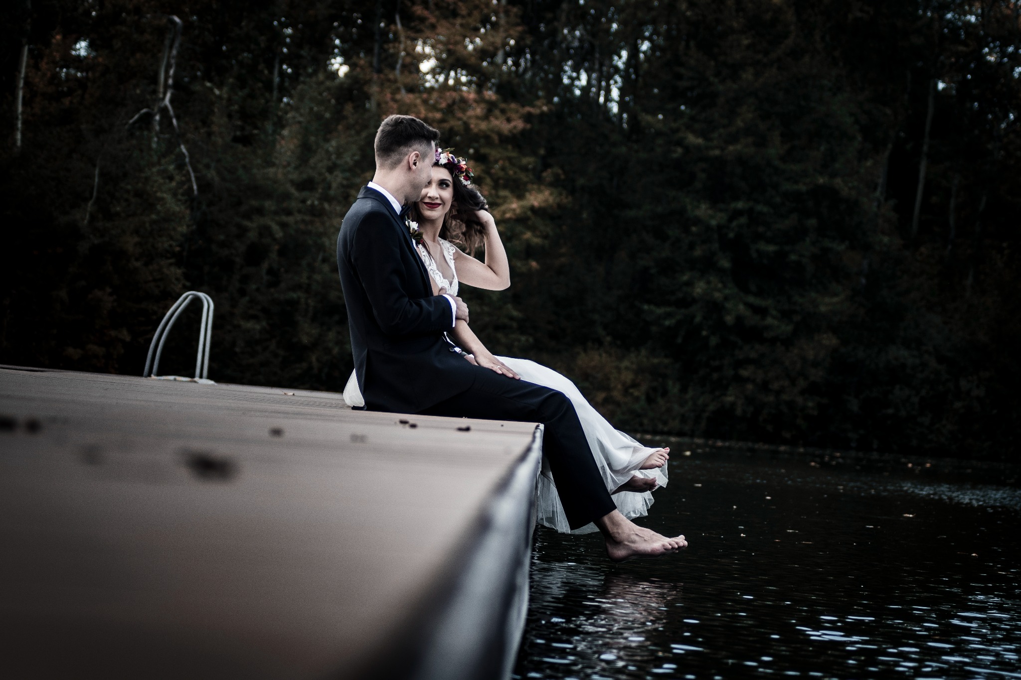 Para młoda siedzi na drewnianym pomoście nad wodą, boso, ona w wianku na głowie, on w czarnym garniturze, w tle ciemny las.