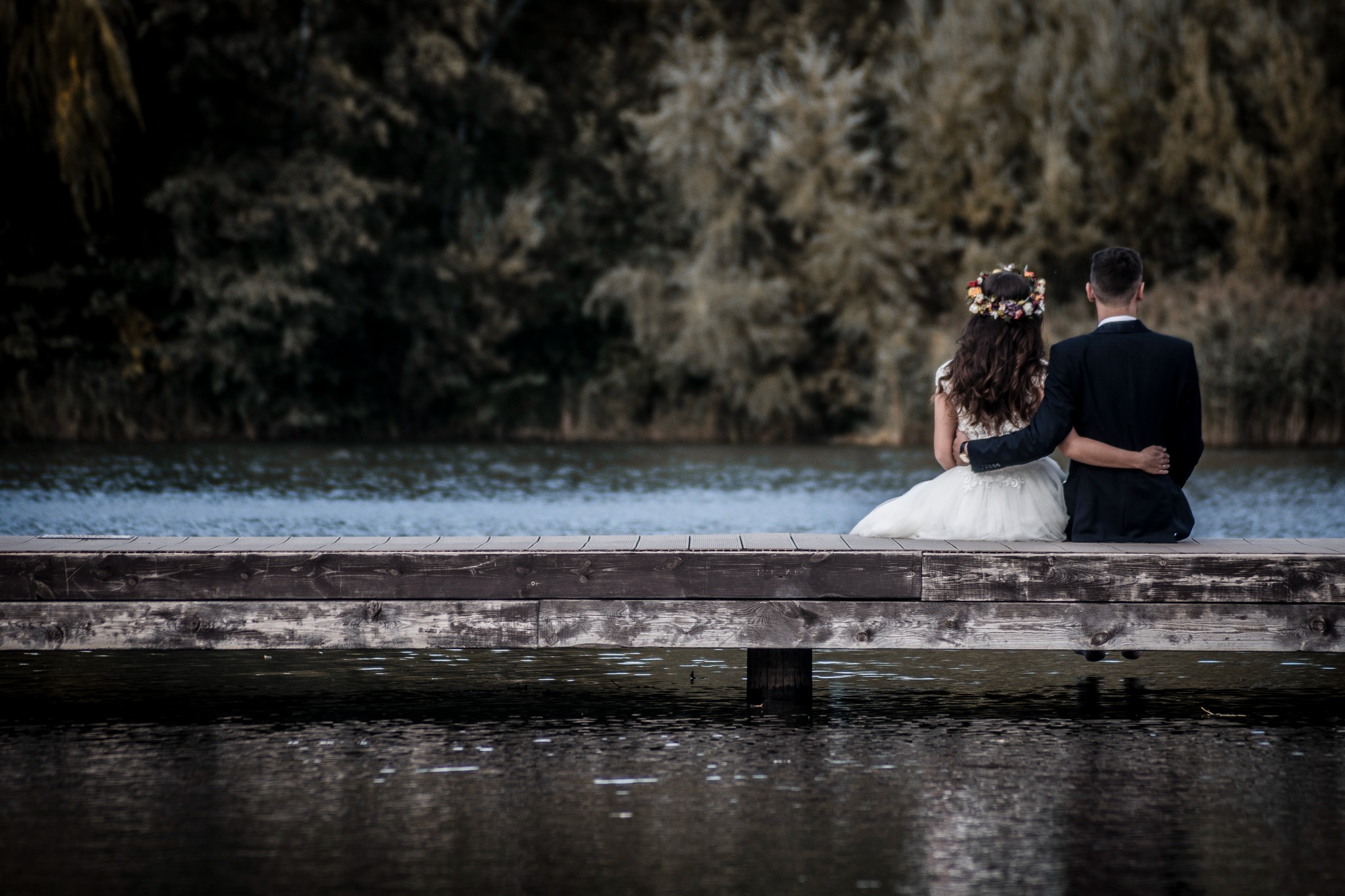 Para młoda siedzi na drewnianym pomoście nad wodą, obejmując się i patrząc na drzewa w tle. Panna młoda ma wianek z kwiatów na głowie.