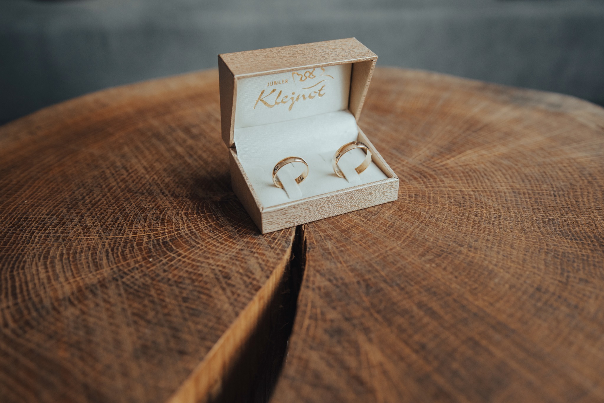 Dwa złote obrączki ślubne w beżowym pudełku jubilera Klejnot, umieszczone na okrągłym drewnianym blacie z wyraźnym usłojeniem i pęknięciem.