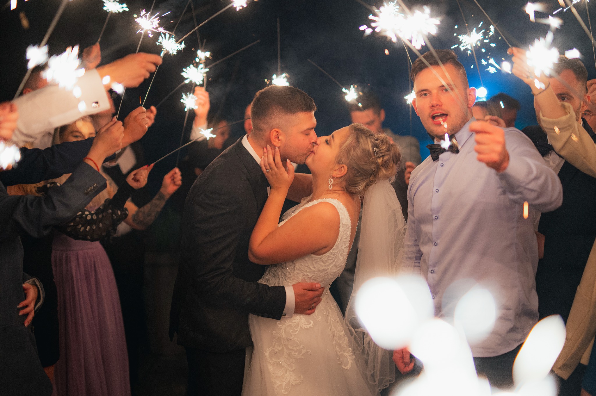 Pocałunek nowożeńców otoczonych gośćmi z zimnymi ogniami, panna młoda w koronkowej sukni i welonie, pan młody w ciemnym garniturze, radosna atmosfera.