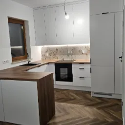 Nowoczesna kuchnia z białymi szafkami, drewnianym blatem i podłogą ułożoną w jodełkę, oświetlona pojedynczą żarówką.