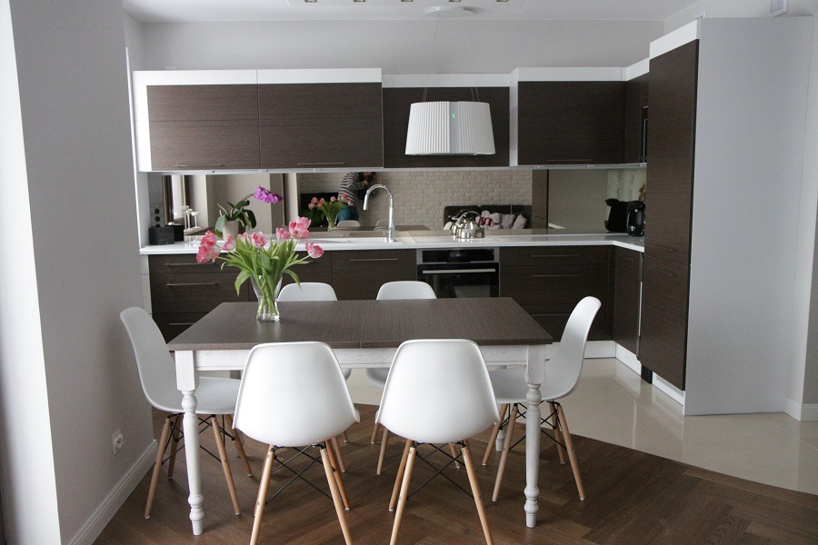 Nowoczesna kuchnia z ciemnymi, drewnopodobnymi frontami szafek, białymi blatami i lustrzanym panelem nad blatem, widoczny stół z białymi krzesłami i bukiet tulipanów.