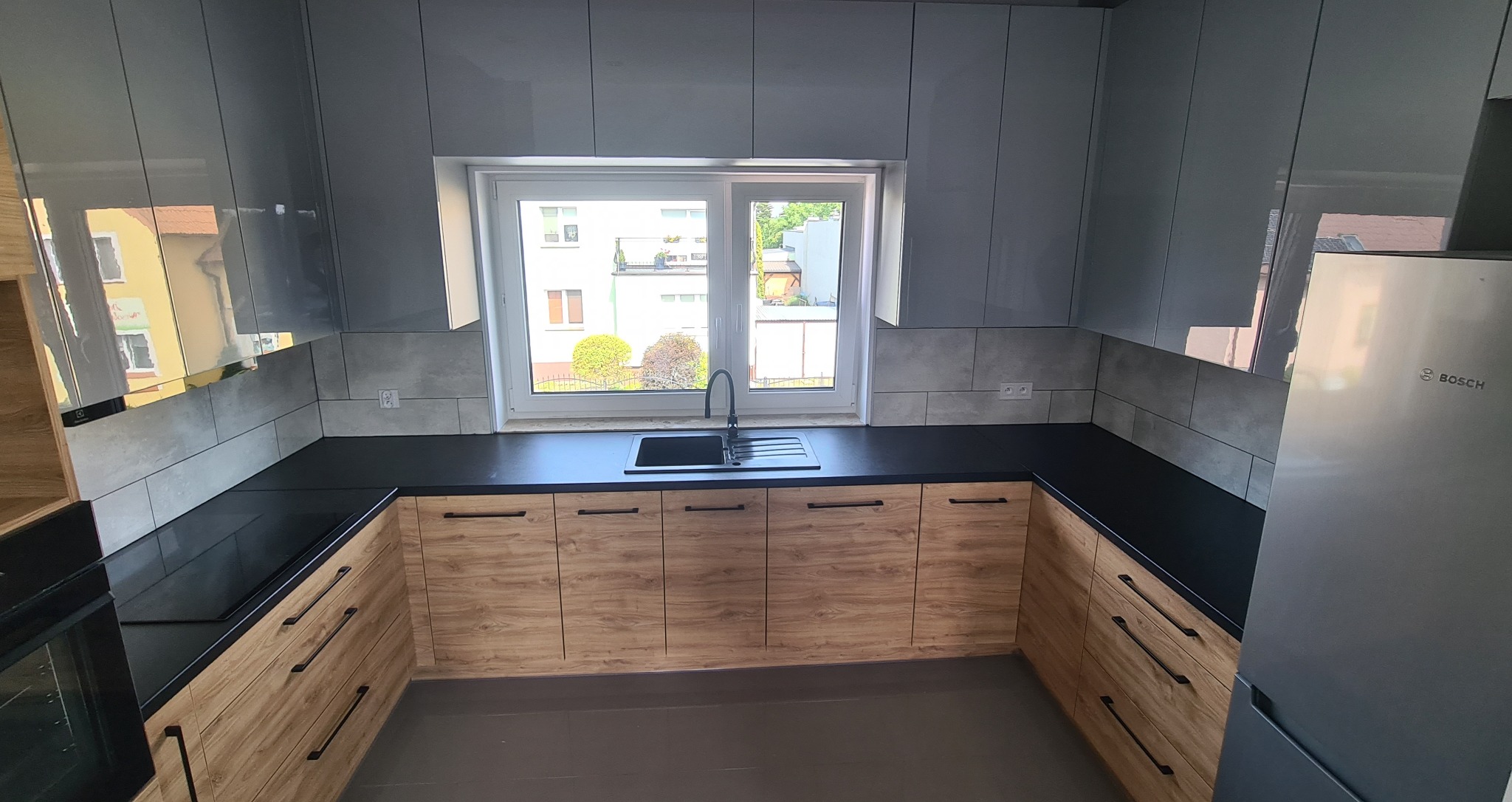 Nowoczesna kuchnia z szarymi, lakierowanymi szafkami górnymi i drewnopodobnymi szafkami dolnymi, czarnym blatem i widokiem z okna na budynki mieszkalne.