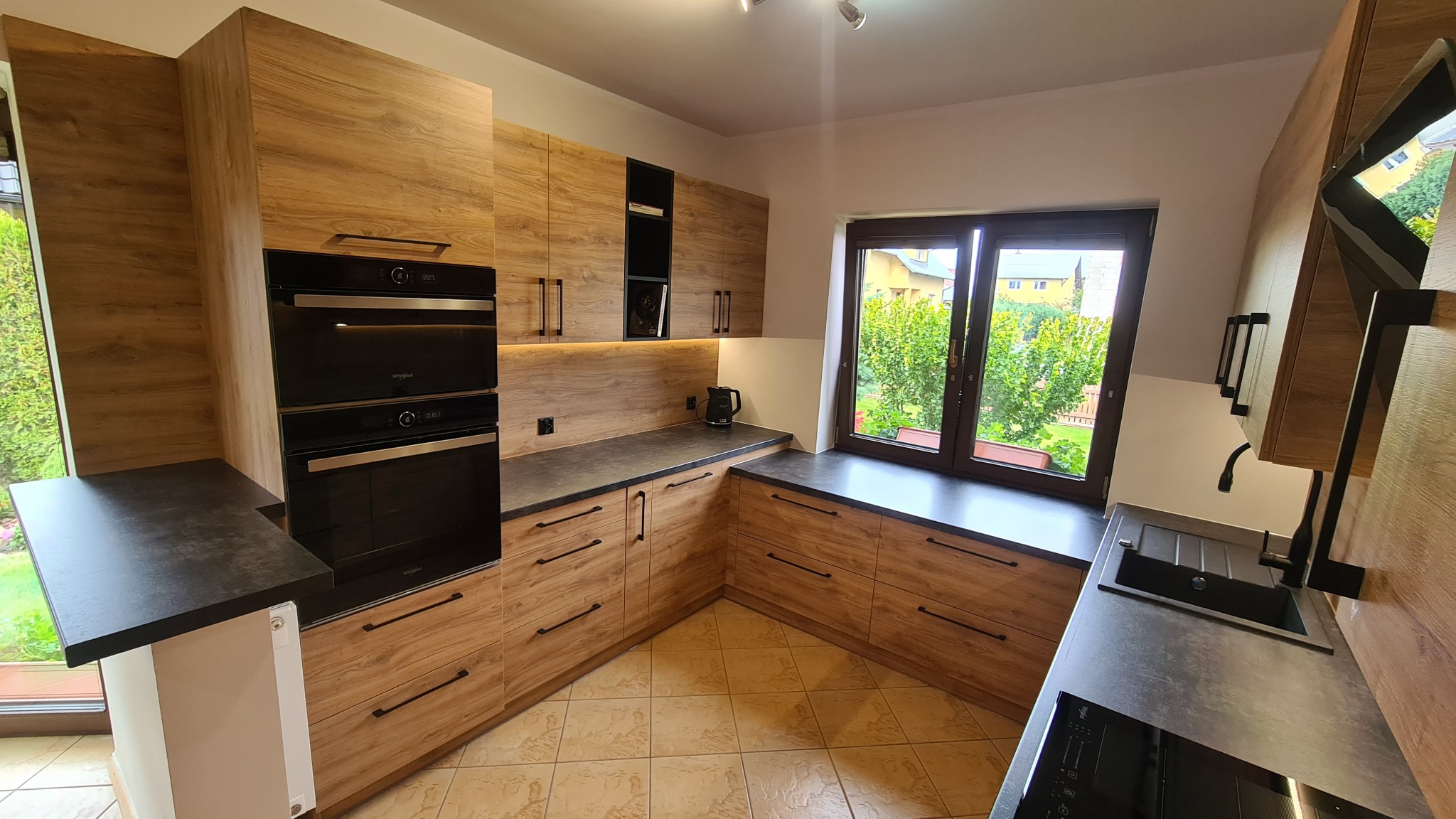Nowoczesna kuchnia z drewnianymi frontami i czarnymi akcentami w Chojnicach, z widokiem na zieleń za oknem, ukazująca zabudowę piekarnika i płytę grzewczą.