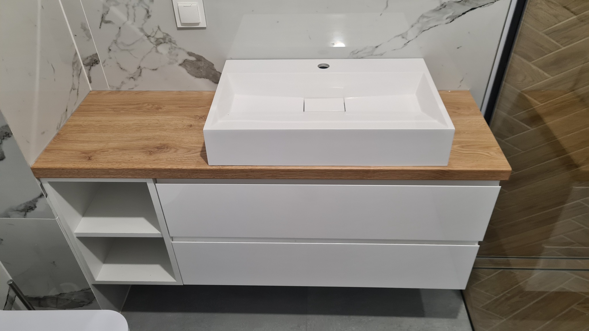 Nowoczesna biała umywalka nablatowa o kwadratowym kształcie, osadzona na szafce z drewnianym blatem o jasnym odcieniu dębu, z widocznymi szufladami i otwartymi półkami, na tle marmurowej ściany...