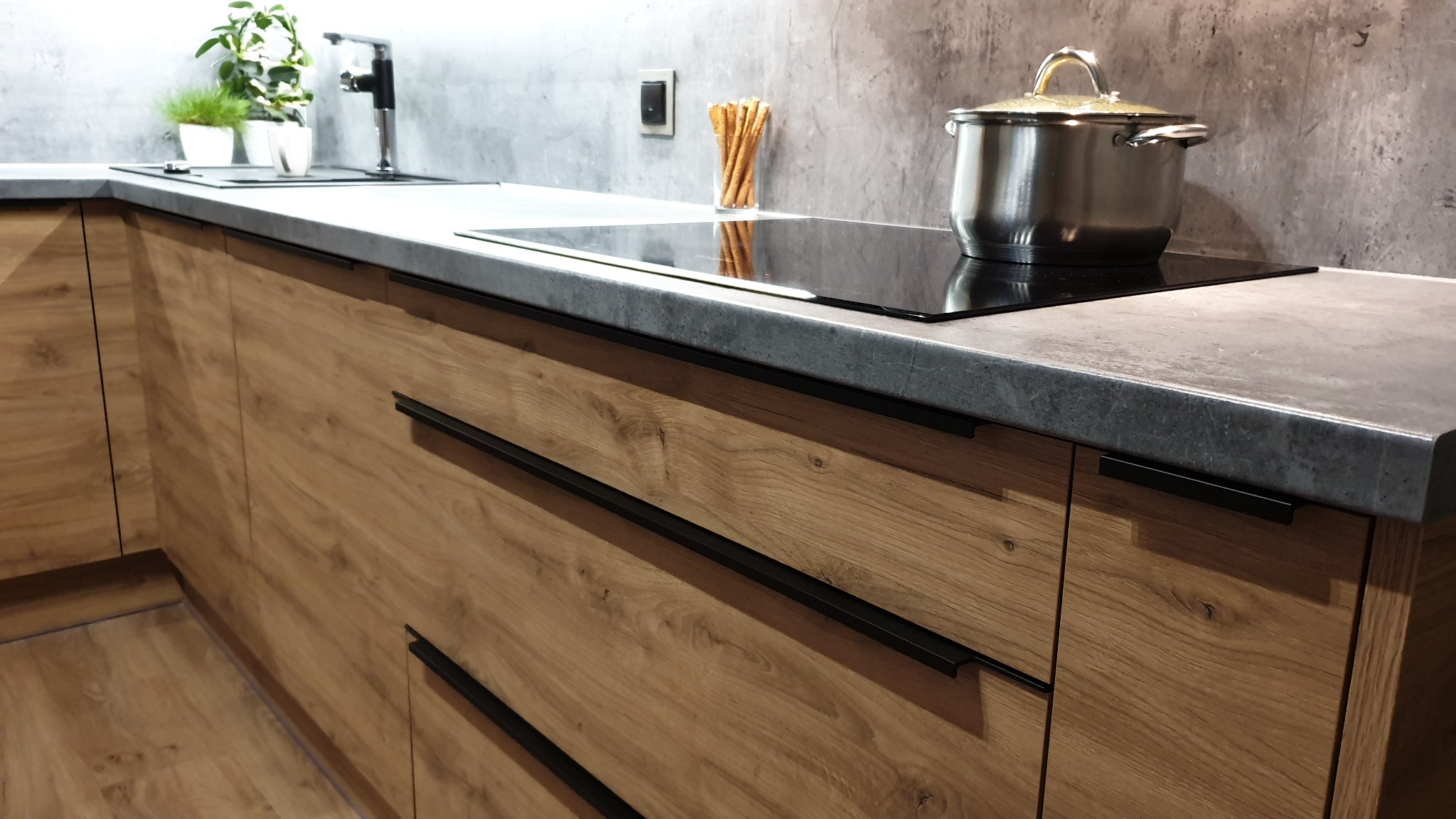 Nowoczesna kuchnia z drewnianymi frontami, szarym blatem i czarnymi uchwytami. Na blacie indukcyjnym stoi garnek, obok w szklance paluszki.