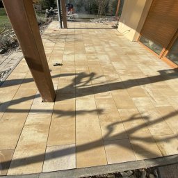 Dawid - Wykończony taras z beżowej kostki brukowej, z drewnianym słupem i cieniem. W tle widoczny pracownik podczas cięcia kostki, co sugeruje trwające prace wykończeniowe.