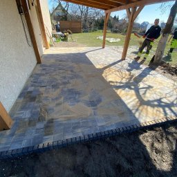 Dawid - Ułożona kostka brukowa pod drewnianą pergolą, widoczni dwaj pracownicy w odblaskowych ubraniach, teren jeszcze niezagospodarowany. Kostka w odcieniach szarości.