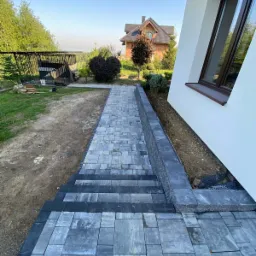 Ukończone schody i ścieżka z szarej kostki brukowej prowadzące wzdłuż białego budynku. Widoczne elementy ogrodu i dom w tle. Kąt ujęcia z dołu.
