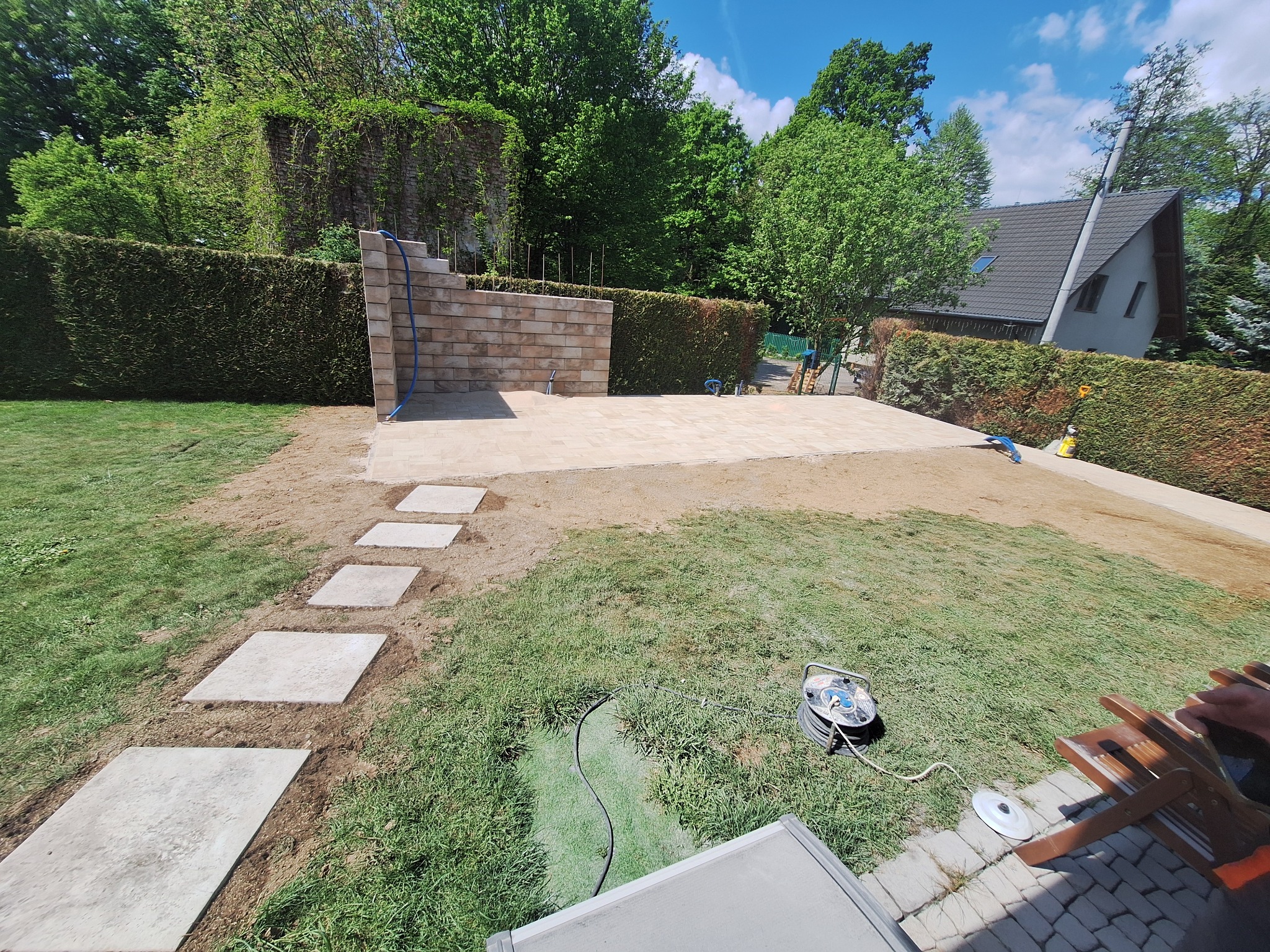 Wykończony taras z jasnej kostki, z murem oporowym z tego samego materiału. Ścieżka z płyt prowadzi przez trawnik. W tle żywopłot i dom.