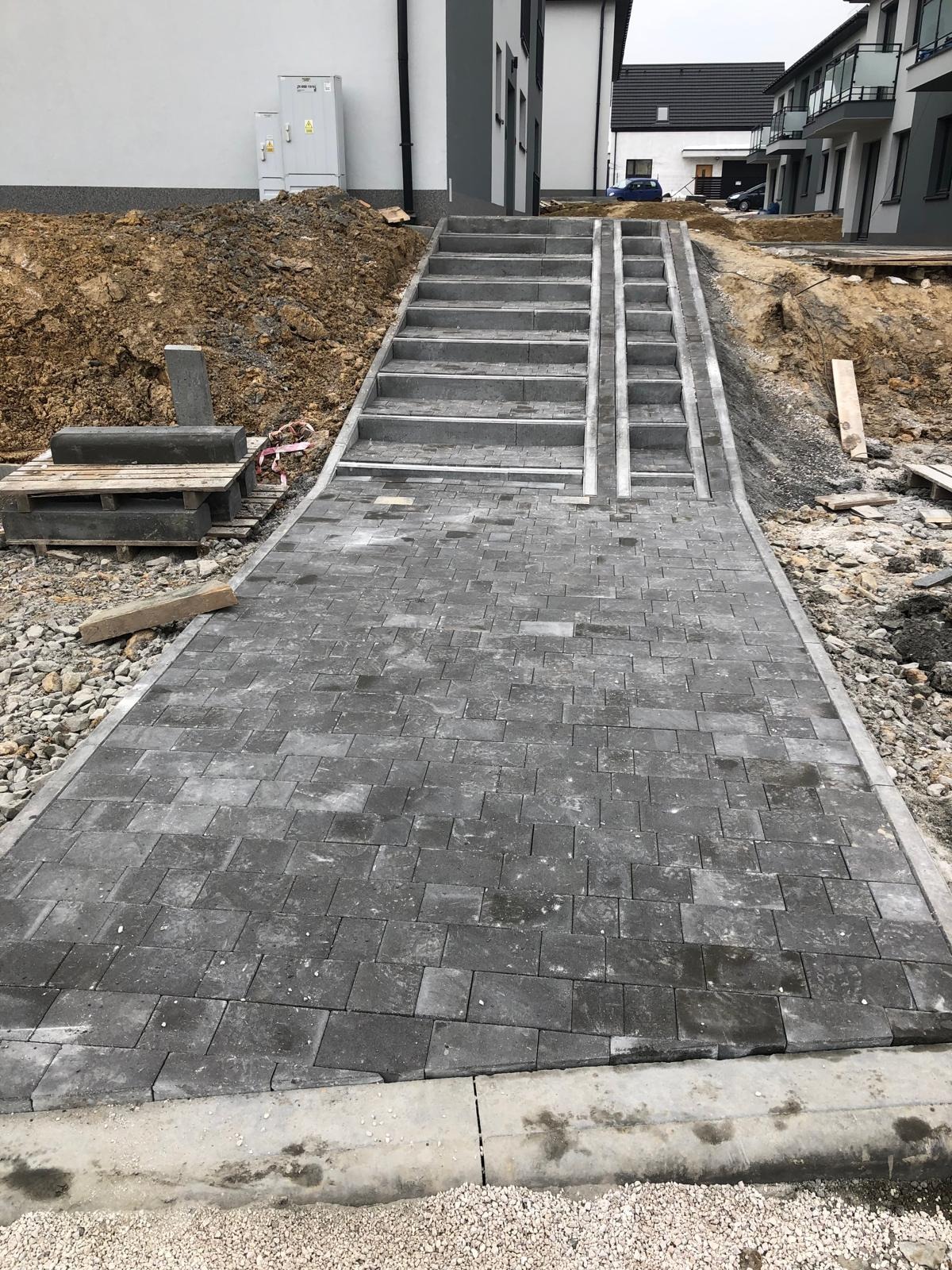 Ułożona, szara kostka brukowa prowadząca do schodów na posesji, z widocznymi elementami palety z kostką. Praca w toku, widoczne pozostałości ziemi i materiałów budowlanych.