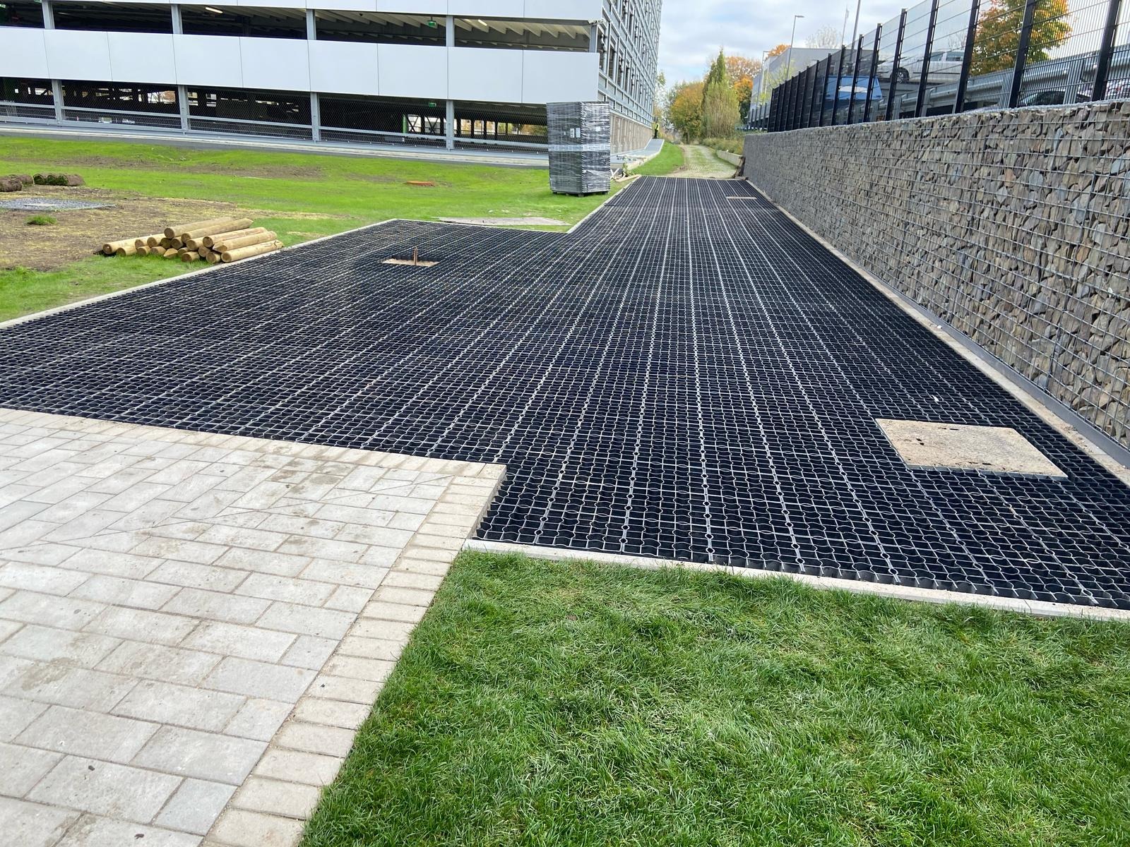 Utwardzona nawierzchnia z kraty trawnikowej, obrzeże z kostki, mur oporowy z kamienia, trawnik i budynek w tle. Widok z perspektywy.