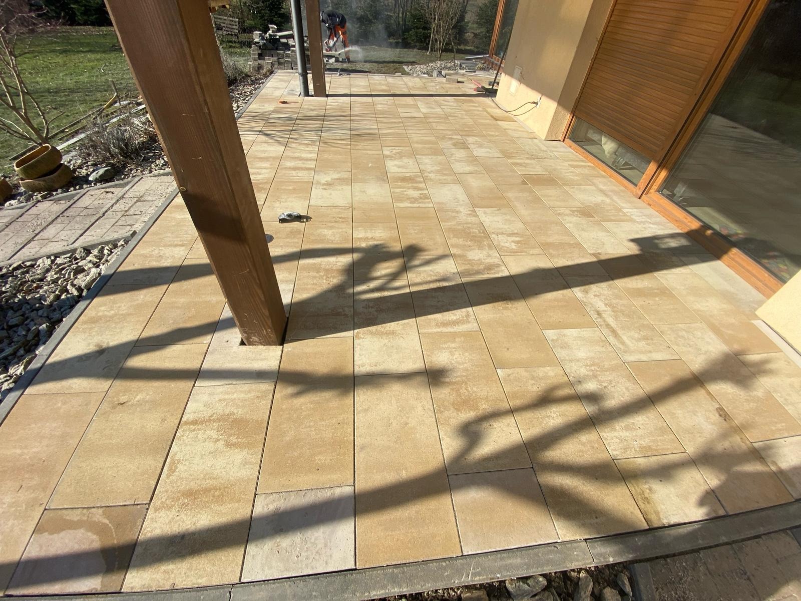 Wykończony taras z beżowej kostki brukowej, z drewnianym słupem i cieniem. W tle widoczny pracownik podczas cięcia kostki, co sugeruje trwające prace wykończeniowe.