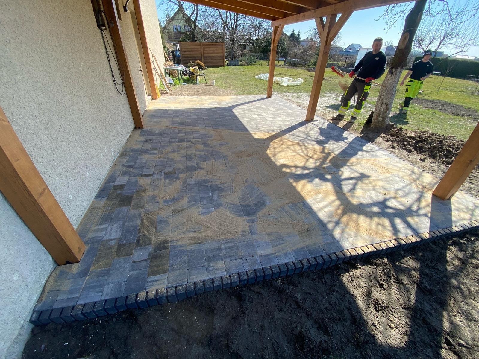 Ułożona kostka brukowa pod drewnianą pergolą, widoczni dwaj pracownicy w odblaskowych ubraniach, teren jeszcze niezagospodarowany. Kostka w odcieniach szarości.