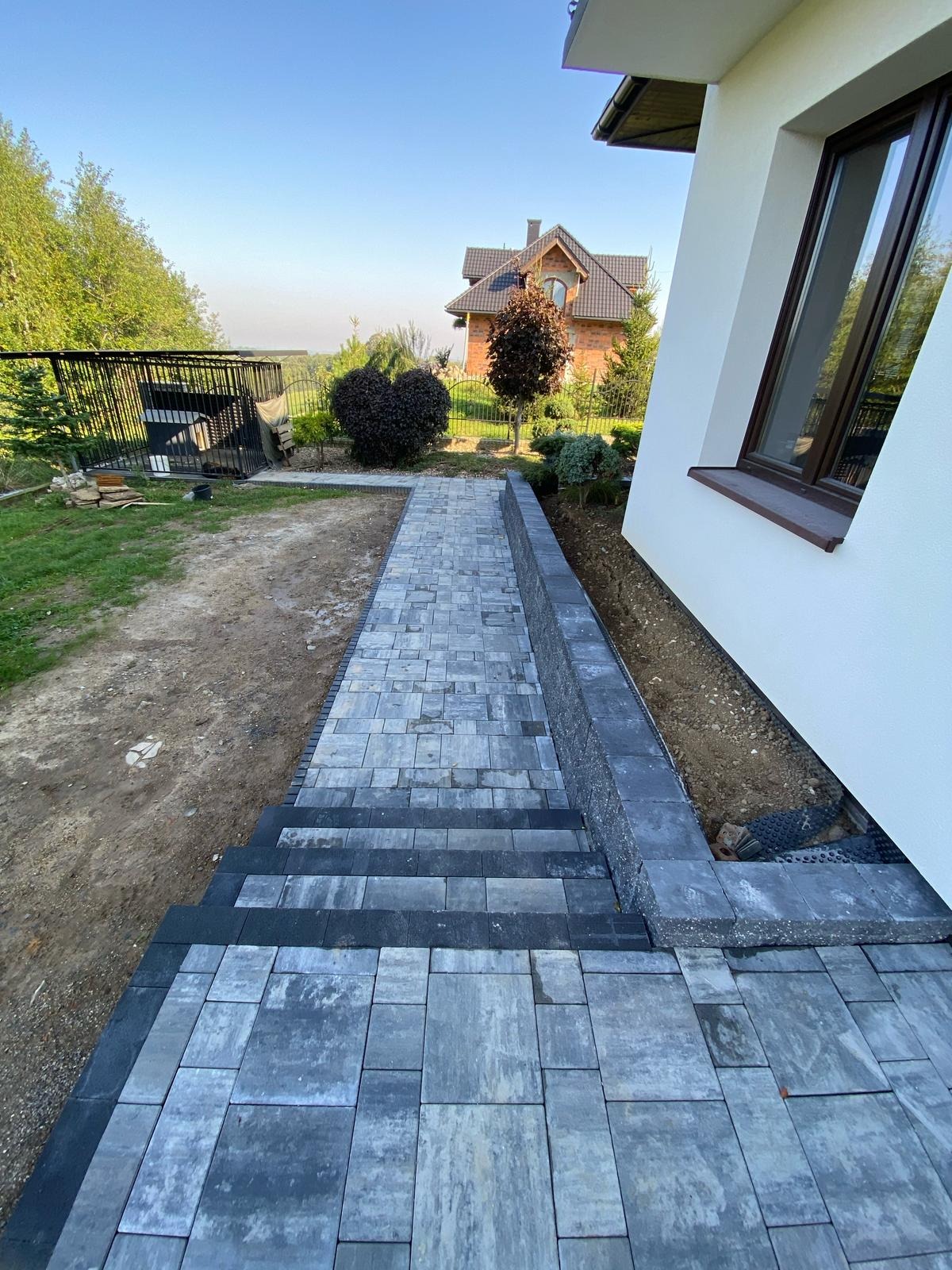 Ukończone schody i ścieżka z szarej kostki brukowej prowadzące wzdłuż białego budynku. Widoczne elementy ogrodu i dom w tle. Kąt ujęcia z dołu.