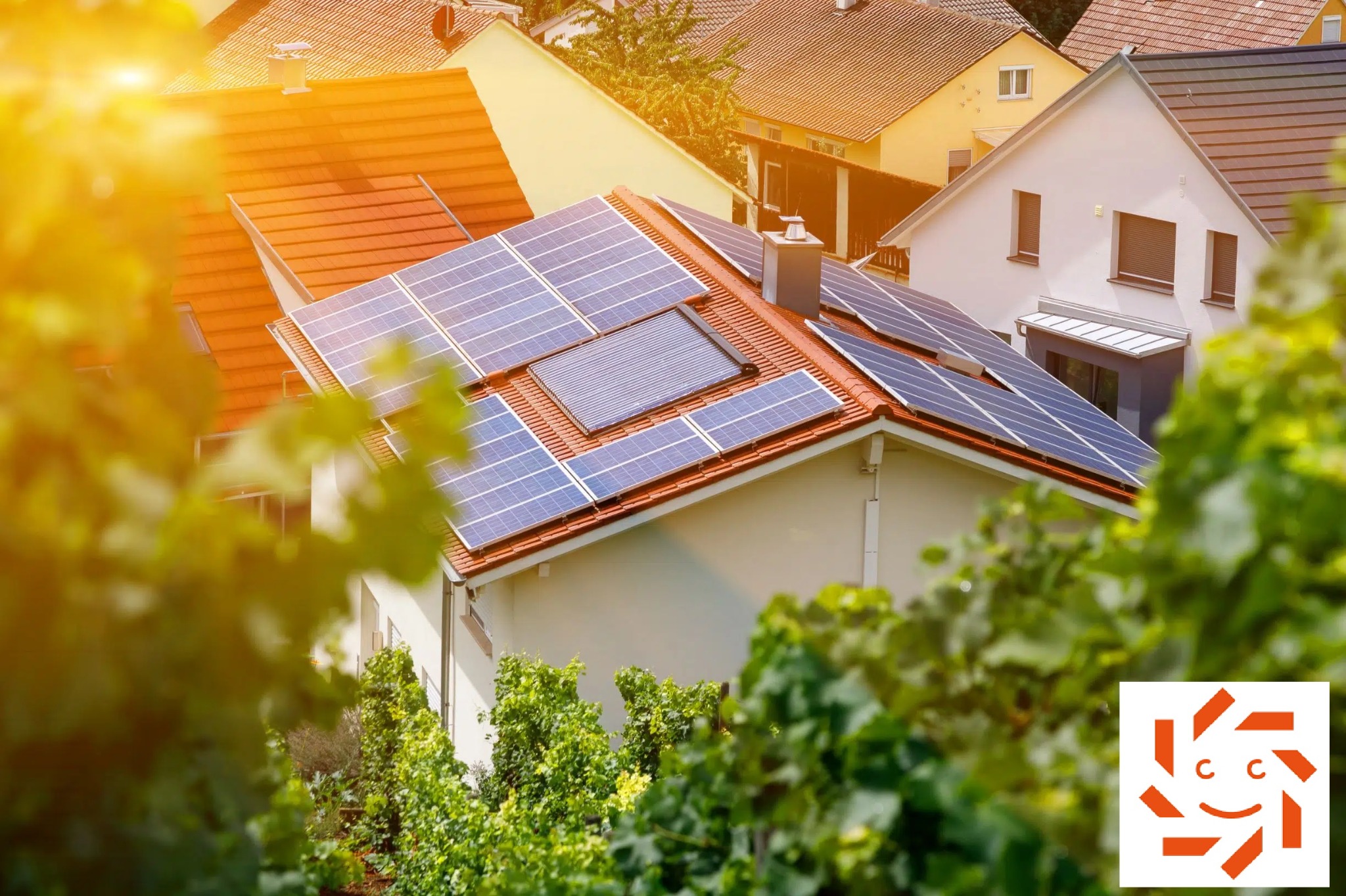 Instalacja fotowoltaiczna na dachu domu jednorodzinnego z czerwoną dachówką, widok zza zieleni, dodatkowo kolektor słoneczny w centralnej części dachu.