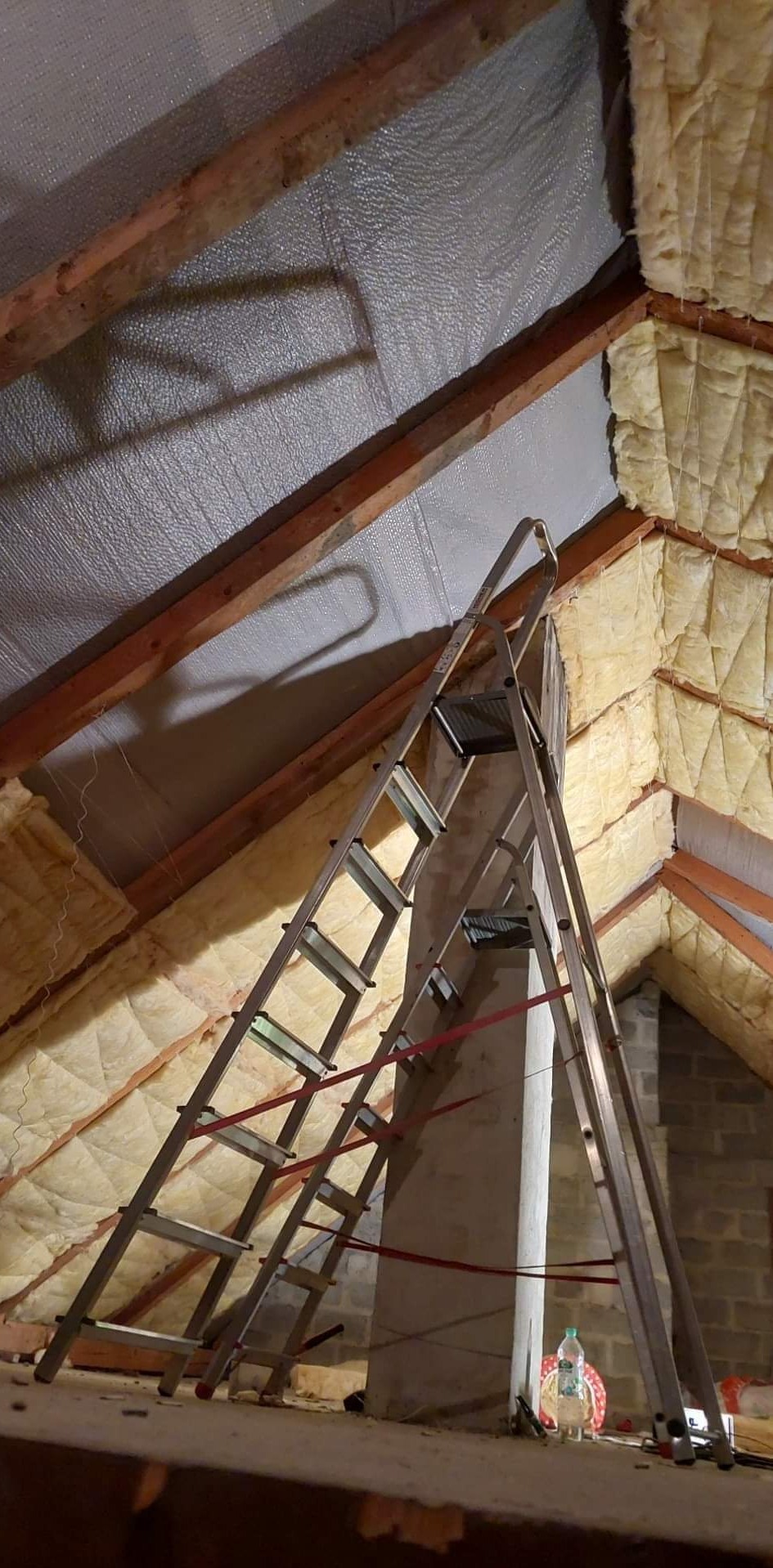 Skośny strych w trakcie prac izolacyjnych, widoczna wełna mineralna między krokwiami, folia aluminiowa, drabina oparta o komin, butelka z wodą na podłodze.