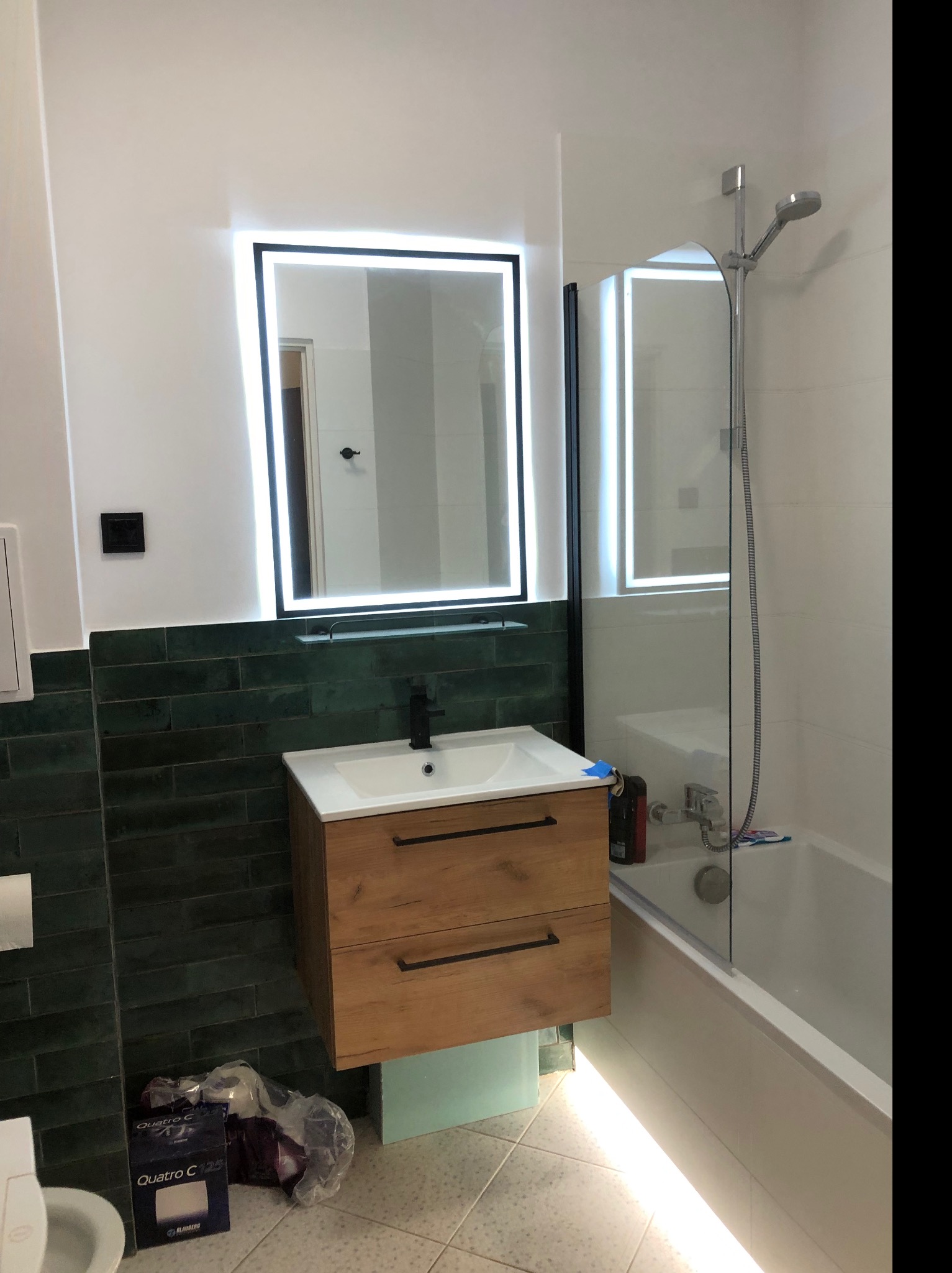 Nowoczesna łazienka z podświetlanym lustrem i szafką pod umywalką z drewnianym frontem, ciemnozielona glazura na ścianie, kabina prysznicowa ze szklaną ścianką, widoczne opakowania po materiałach...