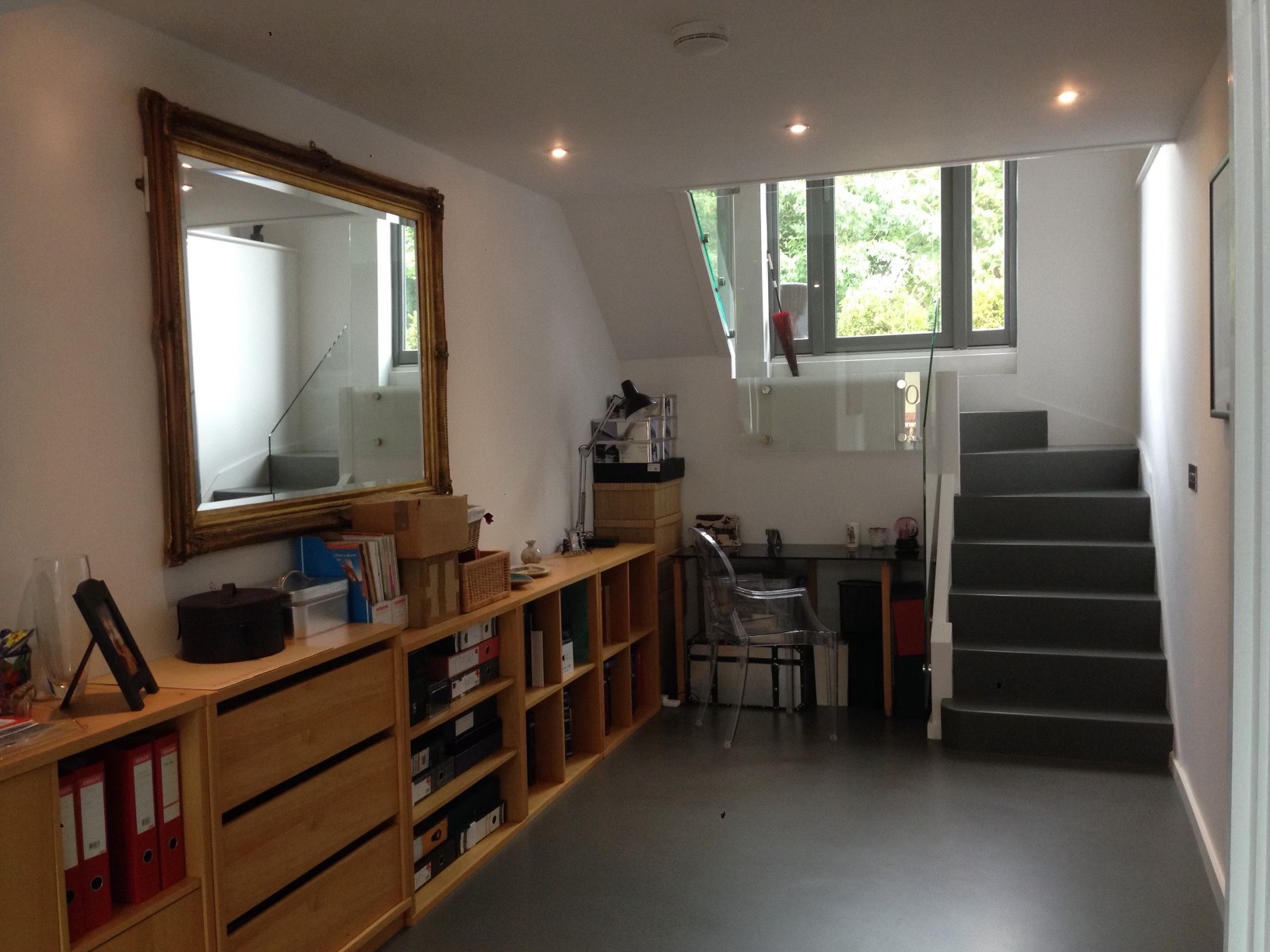 Jasne wnętrze z drewnianymi regałami, dużym lustrem w złotej ramie odbijającym schody, oraz schodami prowadzącymi na górę z przeszkloną balustradą.