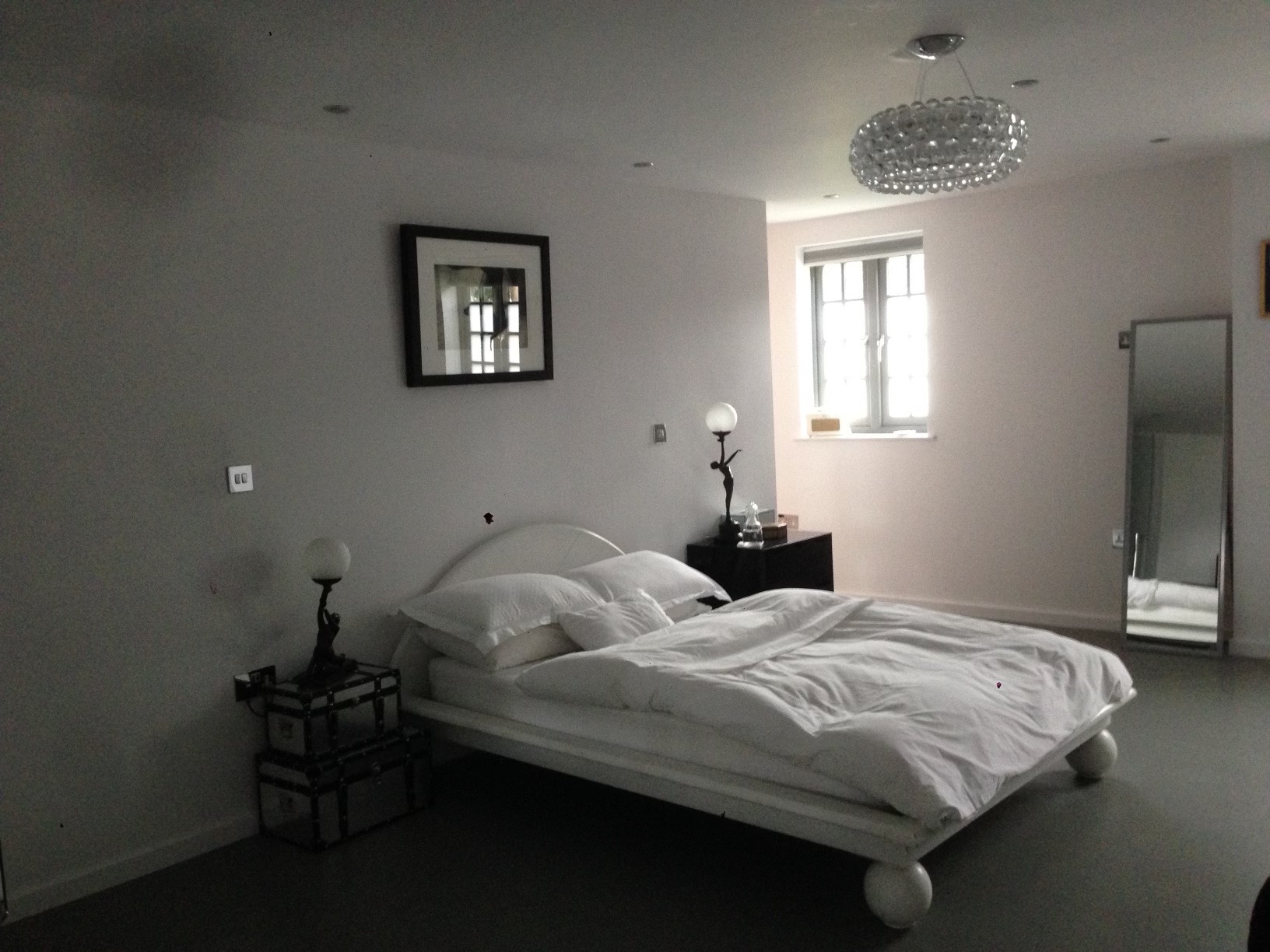 Jasne wnętrze sypialni z białym łóżkiem na okrągłych nogach, obok szafka nocna z lampką i dekoracyjnym elementem, na ścianie obraz w czarnej ramie i kryształowy żyrandol.