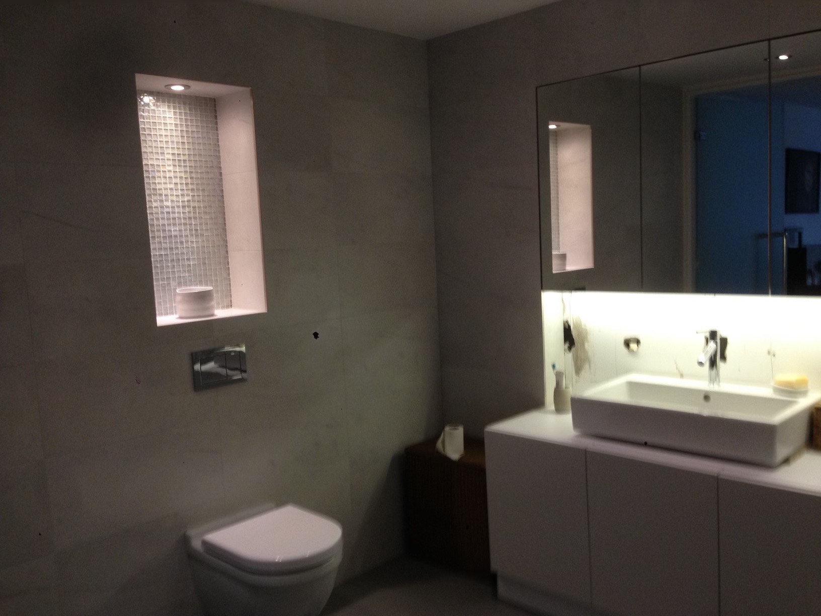 Nowoczesna łazienka z szarą płytką, podświetlaną niszą z mozaiką, białą umywalką nablatową z szafką i toaletą.