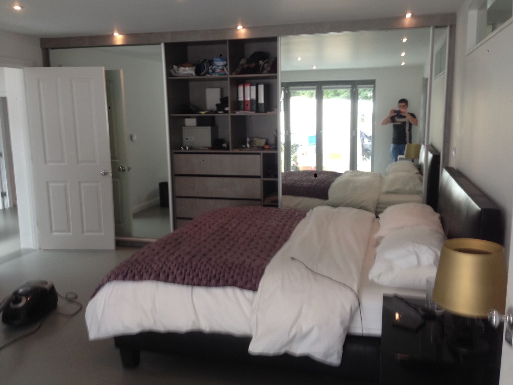 Sypialnia z dużą szafą wnękową z lustrzanymi drzwiami przesuwnymi, widoczna część łóżka z fioletową narzutą i białą pościelą, w lustrze odbija się osoba robiąca zdjęcie telefonem.