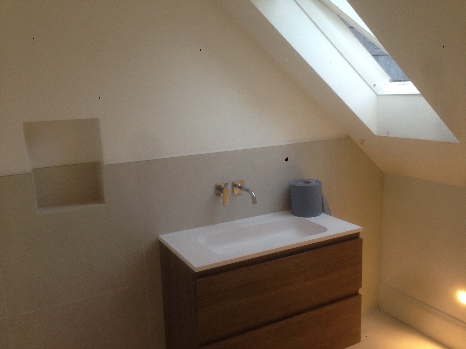 Umywalka nablatowa z drewnianą szafką w łazience na poddaszu, widoczne okno dachowe i wnęka w ścianie wyłożonej płytkami, obok umywalki stoi rolka szarego papieru toaletowego.