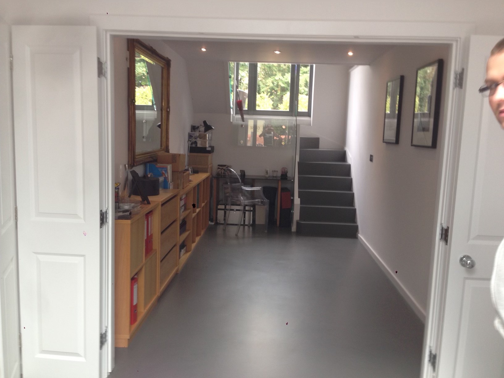 Przestronne wnętrze po remoncie z szarym podłożem, widoczne białe drzwi, drewniany regał z czerwonymi segregatorami, schody prowadzące na górę i okno z widokiem na zieleń.