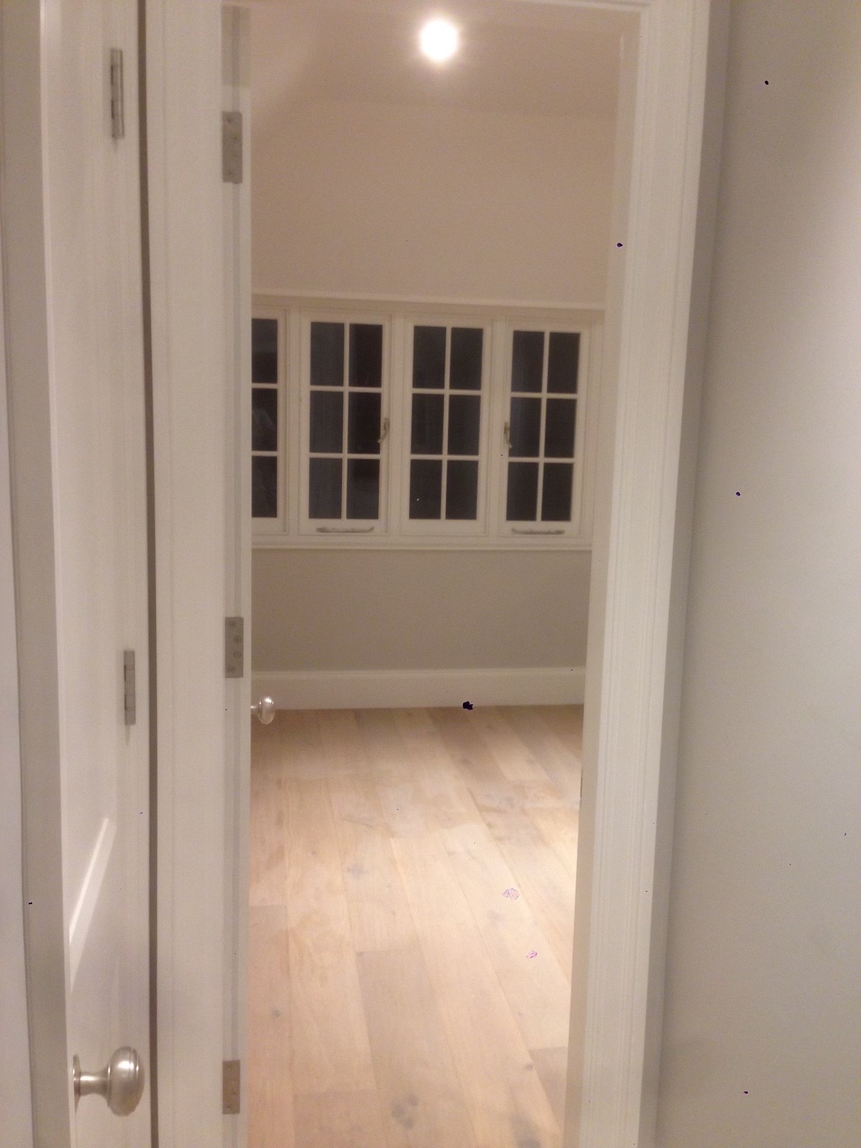 Widok przez otwarte, białe drzwi na jasny pokój z drewnianą podłogą i oknami z białymi ramami, oświetlony górnym światłem.