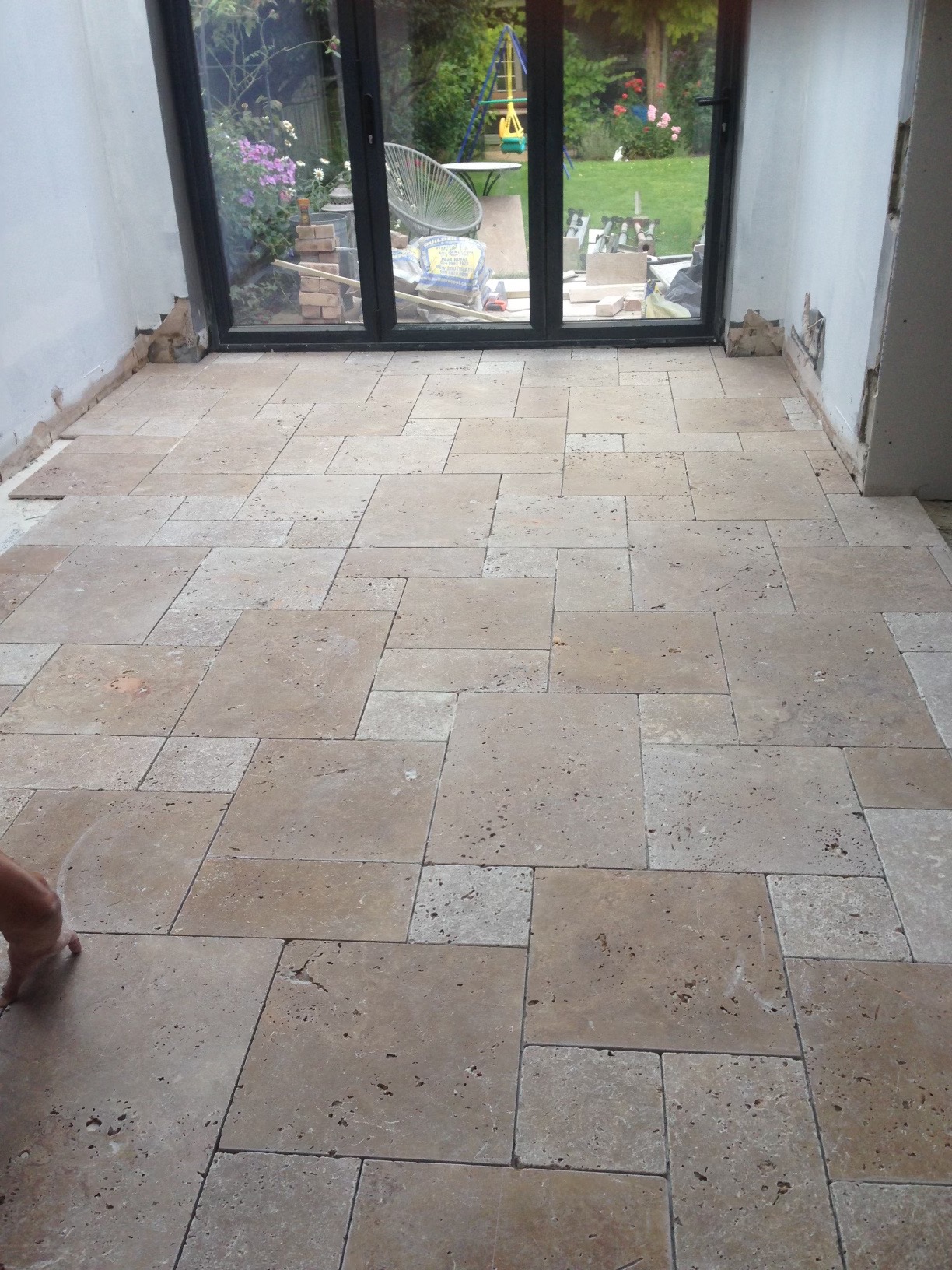 Układanie beżowych, porowatych płytek podłogowych w pomieszczeniu z dużym oknem wychodzącym na ogród, widoczne niedokończone prace przy ścianach.