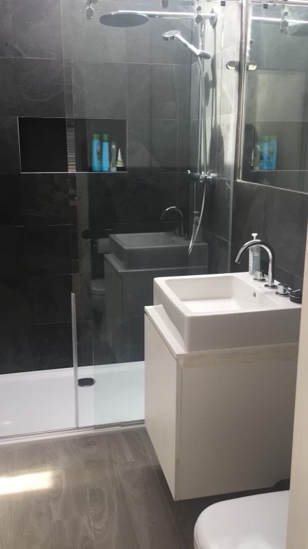 Nowoczesna łazienka z czarnymi płytkami, przeszkloną kabiną prysznicową, białą umywalką nablatową na szafce i drewnopodobną podłogą. W ścianie wnęka z kosmetykami.