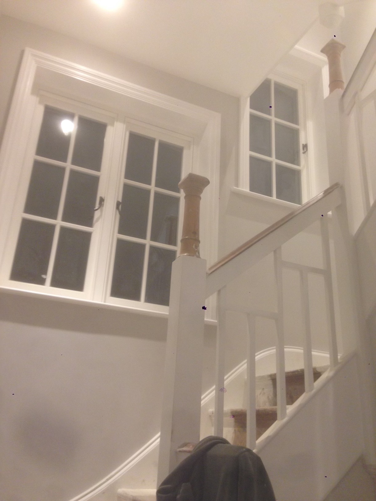 Klatka schodowa z białymi schodami i balustradą, widoczne dwa okna z białymi ramami, ściany w jasnym odcieniu szarości, u dołu kadru fragment ubrania.
