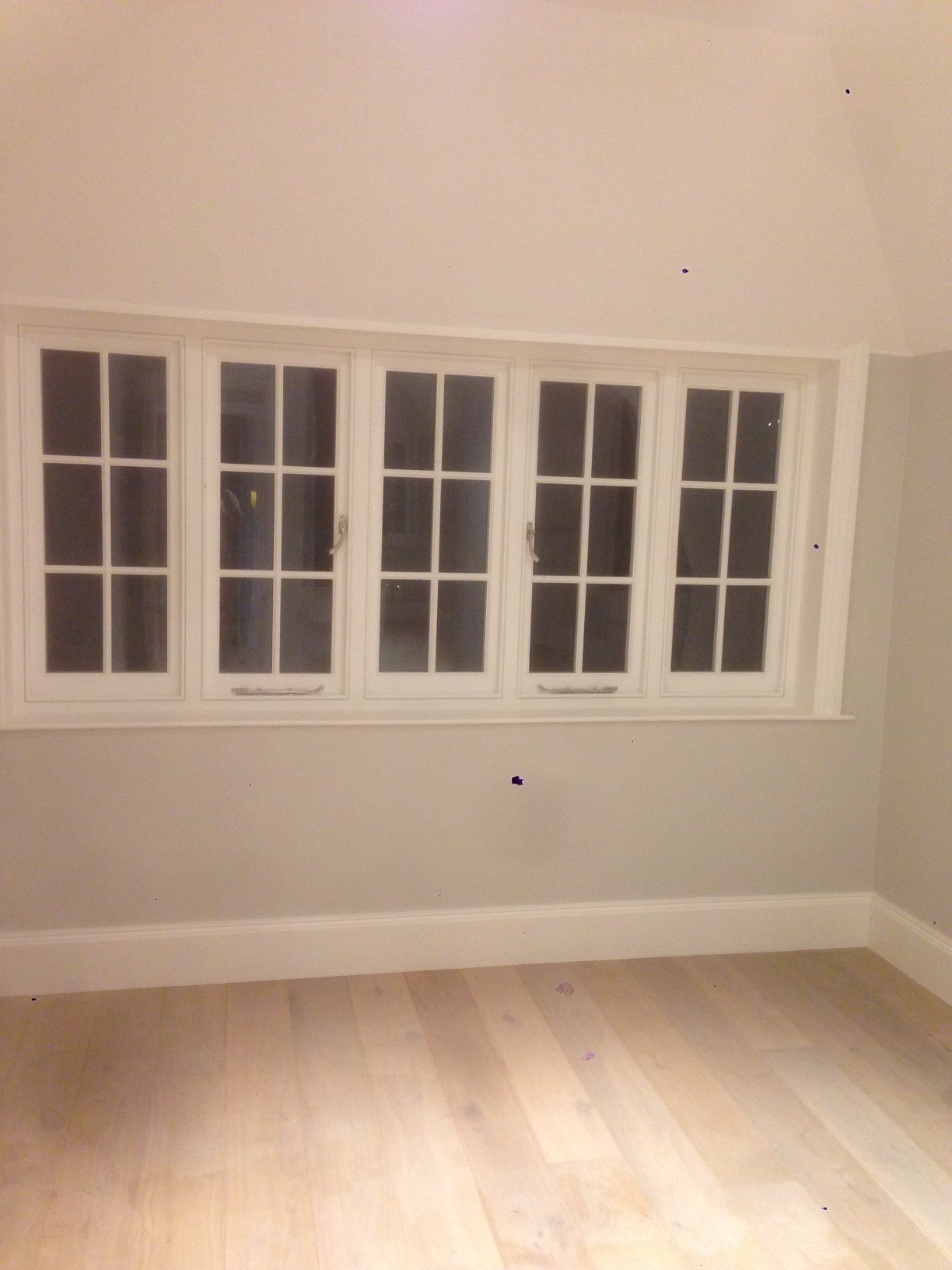 Jasne, drewniane podłogi i ściany w odcieniach szarości w pokoju z pięcioma oknami z białymi ramami i podziałami na mniejsze szybki.