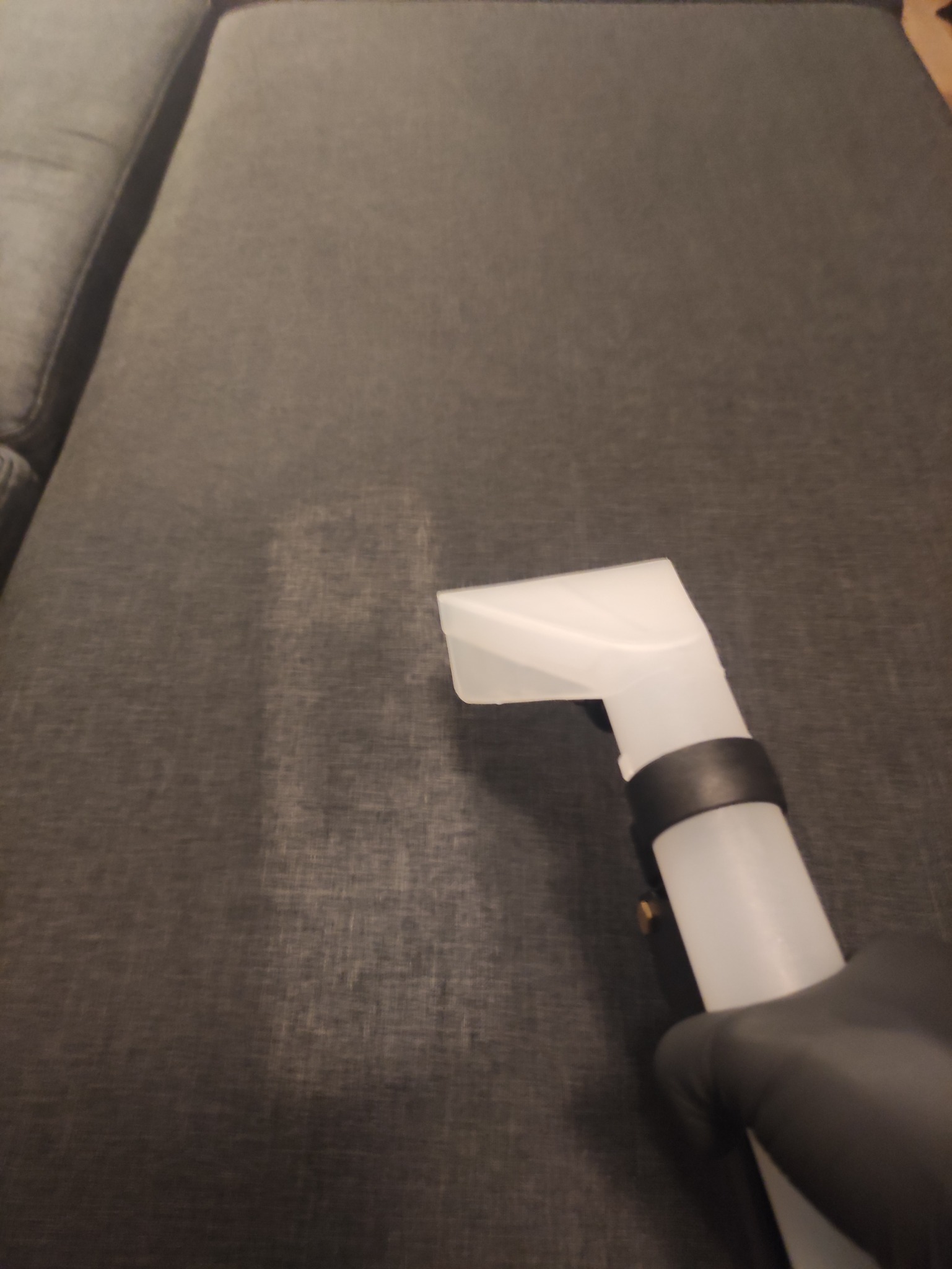 Dysza odkurzacza piorącego na szarej kanapie, widoczny efekt czyszczenia, ręka w czarnej rękawicy.