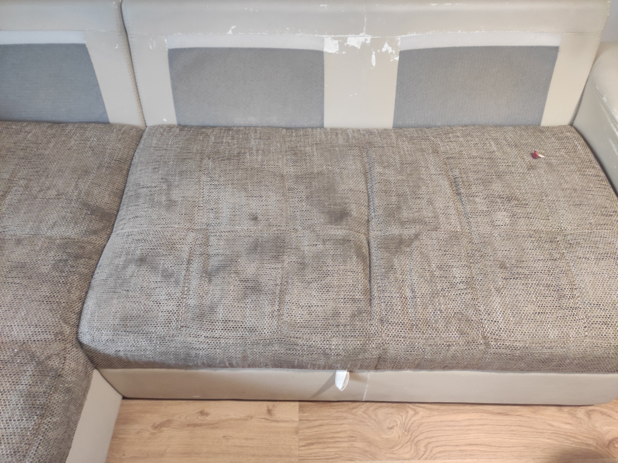 Szaro-beżowa kanapa narożna z widocznymi plamami na siedzisku przed praniem tapicerki, z fragmentem drewnianej podłogi w tle.