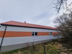 Budynek jednokondygnacyjny z szarą elewacją, poziomym pomarańczowym pasem, czerwonym dachem z dachówki ceramicznej i białymi oknami, widziany z dołu pod kątem, z fragmentem zieleni i drzew po...