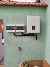 Zewnętrzna instalacja fotowoltaiczna: biała skrzynka inwertera i rozdzielnica elektryczna na szorstkiej, zielonej ścianie budynku z kranem poniżej. Widoczne czarne rury łączące elementy. Częściowo...