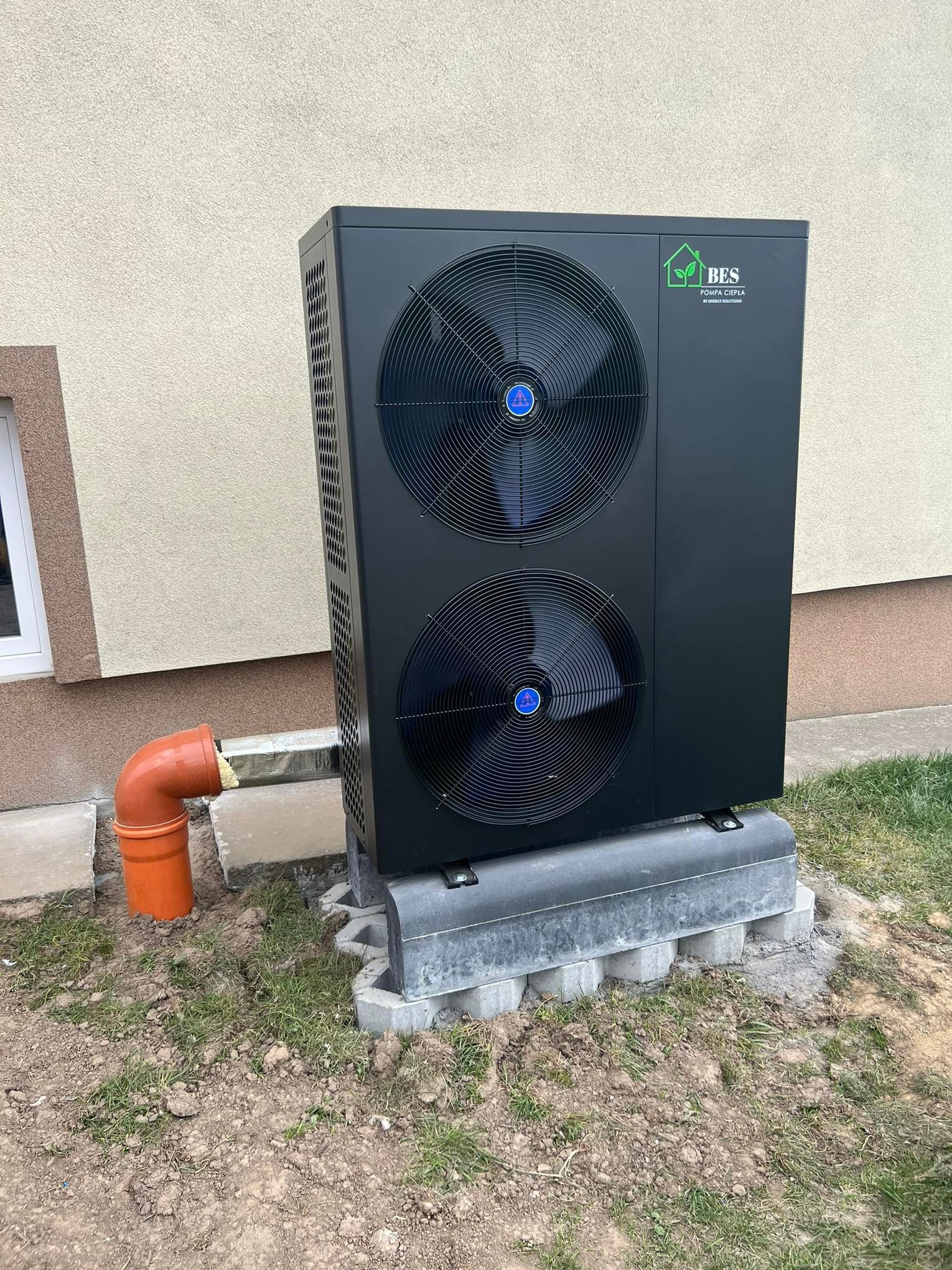 Czarna pompa ciepła monoblok BES z dwoma wentylatorami, zamontowana na betonowych bloczkach przy budynku, z widoczną rurą odpływową koloru pomarańczowego.