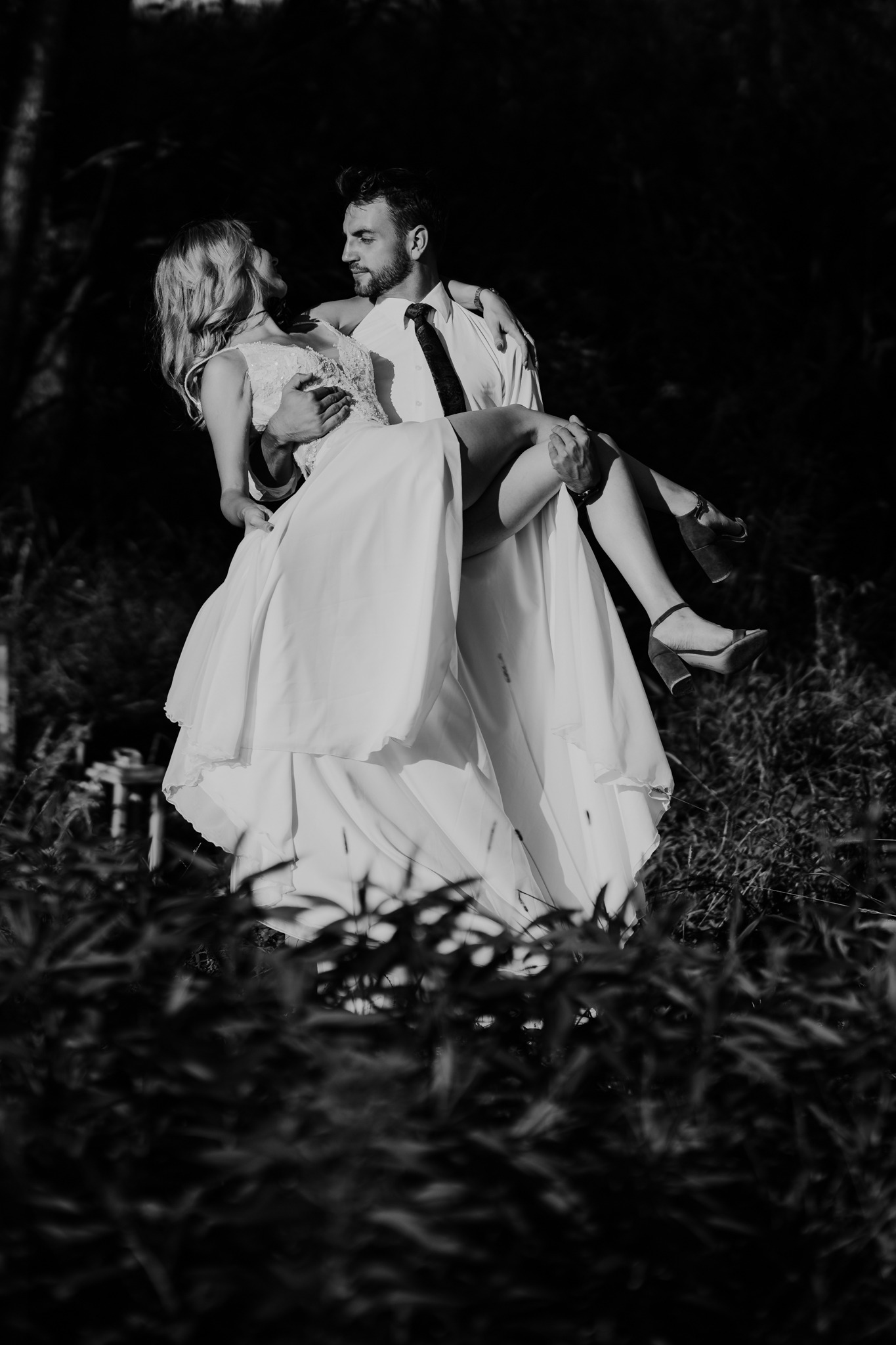 Czarno-białe zdjęcie narzeczonego trzymającego na rękach narzeczoną w sukni ślubnej, stojących w wysokiej trawie.