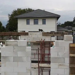Wznoszenie ścian domu z białych bloczków, widoczne rusztowania i butelka wody, w tle gotowy dom i czarny kot spacerujący po ścianie.