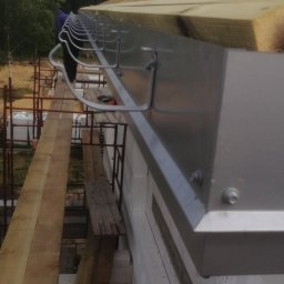 BudArtMagic Mykhailo Zagorodnyi - Montaż metalowej rynny na nowo budowanym domu z białego pustaka, widoczne krokwie z ociepleniem, rusztowanie i fragment sylwetki pracownika.