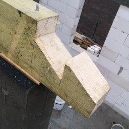 BudArtMagic Mykhailo Zagorodnyi - Fragment konstrukcji domu w trakcie budowy: drewniana belka z wycięciami oparta na szarej kolumnie, w tle białe bloczki ścian z otworem okiennym.