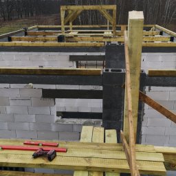 BudArtMagic Mykhailo Zagorodnyi - Wznoszenie ścian z bloczków betonowych na budowie domu, widoczne drewniane szalunki i rusztowania, w tle las bezlistny pod pochmurnym niebem.