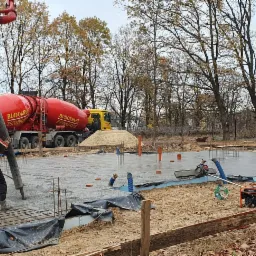 Wylewanie betonu na przygotowany zbrojony fundament pod budowę domu jednorodzinnego, widoczne dwa betonomieszarki z pompami do betonu, pracownicy, zbrojenie, izolacja oraz generator prądu.