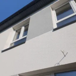 Fragment ocieplonej elewacji domu z dwoma oknami, widoczne różnice w odcieniach szarości tynku oraz czarny orynnowanie pod błękitnym niebem.