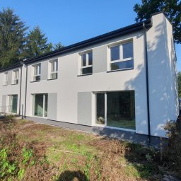 BudArtMagic Mykhailo Zagorodnyi - Nowoczesny, szeregowy budynek mieszkalny z białymi ścianami i szarymi akcentami, duże okna, otoczony zielenią i elementami krajobrazu w trakcie zagospodarowywania.