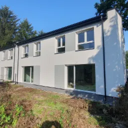 Nowoczesny, szeregowy budynek mieszkalny z białymi ścianami i szarymi akcentami, duże okna, otoczony zielenią i elementami krajobrazu w trakcie zagospodarowywania.